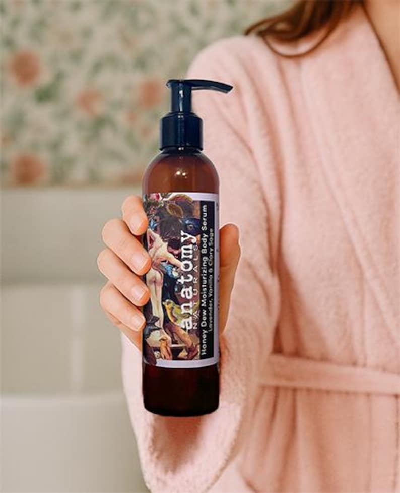 Honey Dew botanical body serum held in a woman’s hand with bathrobe visible, intensive post shower body serum for dry and sensitive skin.