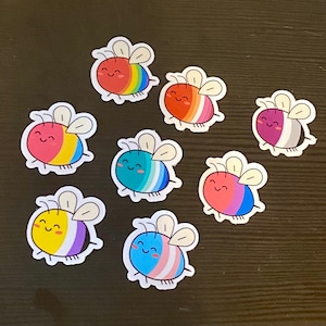 May include: Eight colorful bee stickers with different pride flag designs. The bees are all smiling and have wings. The stickers are on a brown wooden surface.
