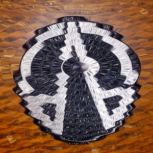 May include: A black and white circular design with concentric circles, resembling a target or bullseye. The design is made of woven materials and has a textured surface.