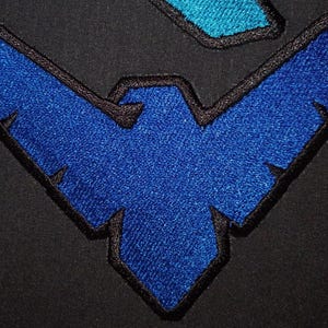 Può includere: Primo piano di un emblema blu ricamato con un contorno nero, che ricorda un uccello o un pipistrello stilizzato, su uno sfondo di tessuto nero. L'emblema ha un aspetto strutturato, con una forma blu più piccola sopra.