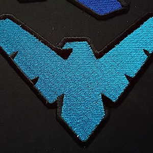 May include: Embroidered patch featuring a stylized blue bird emblem with a black border, set against a black background. The bird design has sharp angles and a textured appearance, suggesting a fabric or thread construction.
