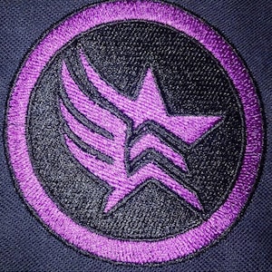 Puede incluir: Parche bordado de color morado con una estrella negra y un diseño de ala estilizado. El parche está rodeado por un círculo morado.