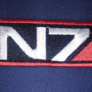 Może przedstawiać: Naszywka haftowana w kolorze czerwonym, czarnym i białym z napisem "N7" w kolorze białym.