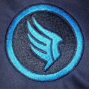 以下が含まれることがあります： 円形のデザインが施された刺繍パッチ。黒い中心と青い縁取りがあります。中央のデザインは、様式化された青い翼の形です。パッチは濃い青色の生地に縫い付けられています。