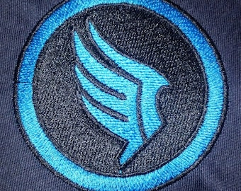 Könnte beinhalten: Gestickter Aufnäher mit kreisförmigem Design. Er hat eine schwarze Mitte und einen blauen Rand. Das mittlere Design ist eine stilisierte blaue Flügelform. Der Aufnäher ist auf einen dunkelblauen Stoff genäht.