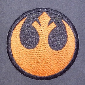 Pode incluir: Um símbolo da Aliança Rebelde bordado em laranja sobre um fundo cinza escuro.