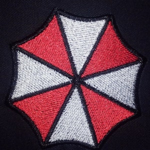 Può includere: Un logo di ombrello ricamato in rosso e bianco con un contorno nero. Il logo è un ombrello stilizzato con otto punte.