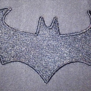 Op de afbeelding: Een zwart geborduurd vleermuis silhouet op een grijze stoffen achtergrond.