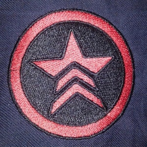 Może przedstawiać: Naszywkę haftowaną w kolorze czerwonym i czarnym z pięcioramienną gwiazdą w środku. Gwiazda ma trzy linie przechodzące przez nią, a naszywka jest otoczona czerwonym kółkiem.