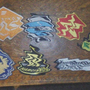 May include: A collection of nine colorful iron-on patches with various designs and text, including "PERK", "GRL", "ENGINSERING", "PUR KLEEN", "NARCISSUS", and "65 32V".