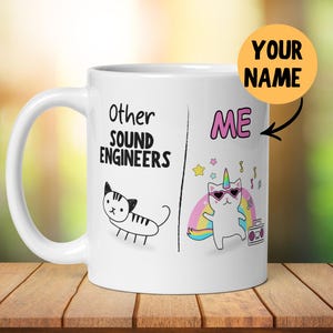 May include: White ceramic mug with a black and white illustration of a cat on the left side and the text "Other Sound Engineers". On the right side is a colorful illustration of a cat with a unicorn horn and sunglasses, with the text "ME" above it. The text "YOUR NAME" is written in a yellow circle on the right side of the mug.