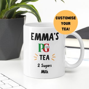 Könnte beinhalten: Weiße Keramiktasse mit schwarzem Text, der "Emma's PG Tips Tea 2 Sugars Milk" lautet. Die Tasse hat einen schwarzen Henkel und steht auf einer weißen Oberfläche.