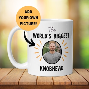 Könnte beinhalten: Eine weiße Keramiktasse mit einem schwarzen und gelben Design. Die Tasse trägt den Schriftzug "The World's Biggest Knobhead" und hat einen Kreis mit einem Platzhalter für ein Foto. Der Text "Add Your Own Picture!" befindet sich in einem gelben Kreis.