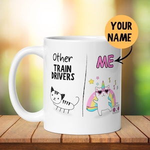 Pode incluir: Caneca de café de cerâmica branca com uma ilustração em preto e branco de um gato no lado esquerdo e um gato unicórnio no lado direito. O texto "Other Train Drivers" está no lado esquerdo e "ME" está no lado direito. O texto "YOUR NAME" está em um círculo amarelo.