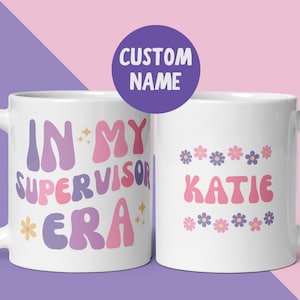 Könnte beinhalten: Zwei weiße Keramiktassen mit buntem Text und Blumengrafiken. Die linke Tasse zeigt den Text IN MY SUPERVISOR ERA in wellenförmigen rosa, lila und gelben Buchstaben mit Sternakzenten. Die rechte Tasse zeigt den Namen KATIE mit Blumenmotiven.