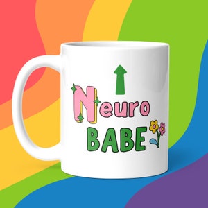 Op de afbeelding: Een witte keramische mok met een groene pijl die omhoog wijst en de tekst "Neuro Babe" in roze en groene letters. De letters zijn zwart omrand en hebben groene sterren eromheen. Rechts van de tekst is een kleine groene bloem met een roze hartje.