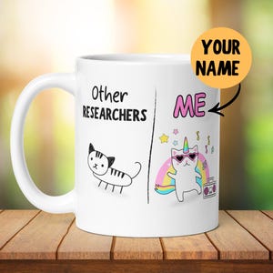 May include: A white ceramic mug featuring a split-screen graphic. The left side shows a simple black line drawing of a cat labelled Other Researchers. The right side shows a colourful cartoon cat unicorn wearing heart sunglasses labelled ME.