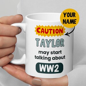 Op de afbeelding: Witte keramische mok met de tekst "CAUTION TAYLOR may start talking about WW2". De mok heeft een geel burst-ontwerp met rode tekst en een groen tekstvak. Er is ook een gele cirkel met de woorden "YOUR NAME".