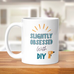 Op de afbeelding: Witte keramische mok met een blauw en oranje ontwerp. De mok heeft de tekst "Slightly Obsessed with DIY" en een cartoon afbeelding van een boormachine.
