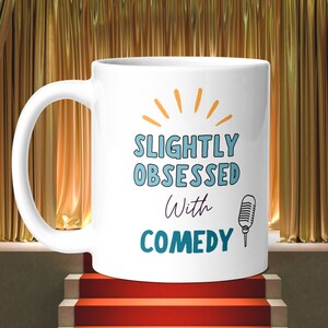 Puede incluir: Taza de café de cerámica blanca con un gráfico de micrófono en blanco y negro y el texto "Slightly Obsessed with Comedy" en azul y morado.