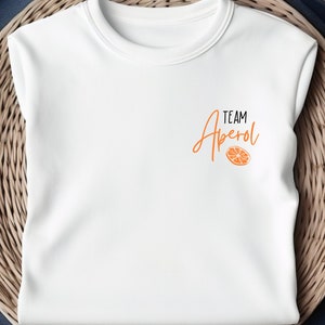 Könnte beinhalten: Weißes T-Shirt mit dem Text "TEAM Aperol" in orangefarbener Schrift mit einer kleinen Grafik einer Orangenscheibe.