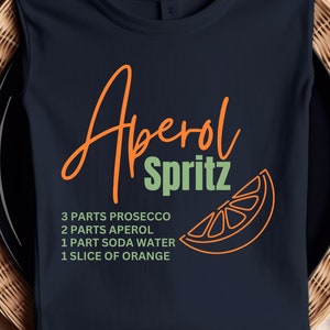 Könnte beinhalten: Ein marineblaues T-Shirt mit dem Text "Aperol Spritz" in Orange und Grün. Der Text wird von einer Grafik einer Orangenscheibe begleitet. Das Rezept für das Getränk ist unter dem Text aufgelistet: 3 Teile Prosecco, 2 Teile Aperol, 1 Teil Sodawasser, 1 Scheibe Orange.