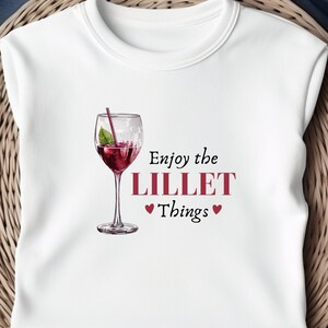 Könnte beinhalten: Weißes Sweatshirt mit Rundhalsausschnitt und einer Cocktail-Illustration in einem Weinglas. Der Text "Enjoy the LILLET Things" ist in Burgunderrot gedruckt, mit zwei Herzsymbolen. Das Design ist mittig auf der Vorderseite des Shirts platziert.