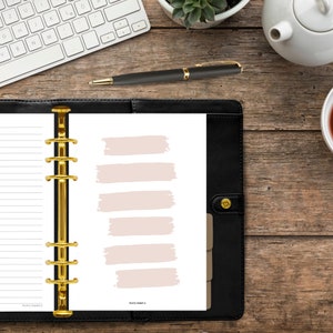 Könnte beinhalten: Ein schwarzer Leder-Planer mit goldenen Ringen und einer weißen Seite mit sechs leeren rosa Pinselstrich-Kästchen. Der Text "Kerry Smith" befindet sich am unteren Rand der Seite.