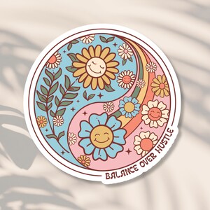 May include: A sticker with a yin yang symbol featuring two smiling suns surrounded by flowers and a rainbow. The text "BALANCE OVER HUSTLE" is written around the symbol.