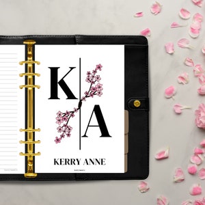 May include: A black leather planner with gold rings and a white page insert. The page has a black and white monogram "KA" with a pink floral design. The text "Kerry Anne" is printed below the monogram.