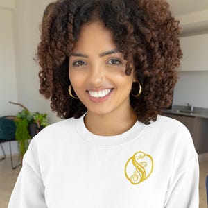 May include: A white sweatshirt with a gold embroidered design of a stylized letter "S" surrounded by floral elements.