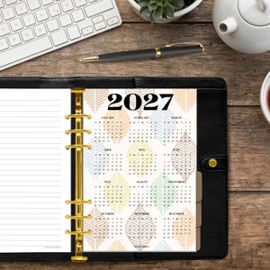 May include: A black leather binder with a gold six-ring mechanism containing a 2027 calendar insert. The calendar page features a repeating leaf pattern in muted colors with each month from January to December clearly displayed.