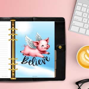 Puede incluir: Un planificador negro con detalles dorados y una inserción azul. La inserción muestra un cerdo rosa con alas blancas, la palabra "Believe" y un fondo de cielo azul. Un teclado blanco y una taza de café están en el fondo.