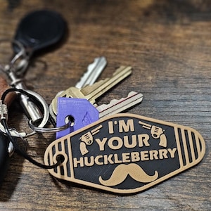 May include: A brown leather keychain with the text "Best Dad Ever" attached to a silver key ring with a black car key fob, a purple key fob, and a wooden keychain with the text "I'm Your Huckleberry" and a brown mustache.