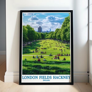 May include: A vintage-style poster with a black frame featuring a scenic view of London Fields Hackney, England. The poster shows a green park with people relaxing on the grass and a cityscape in the background.
