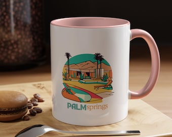Op de afbeelding: Witte keramische mok met een roze rand en handvat. De mok heeft een retro-illustratie van een huis met een zwembad en palmbomen. De tekst "PALMSPRINGS" is in zwart onder de illustratie gedrukt.