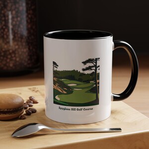 May include: White ceramic coffee mug with a black rim and handle. The mug features a green and brown illustration of a golf course with the text "Spyglass Hill Golf Course".