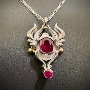 May include: A silver pendant necklace with a large ruby gemstone set in a detailed silver and gold design. The pendant features intricate details and two smaller ruby gemstones.