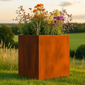 Puede incluir: Una jardinera metálica grande, cuadrada y de color óxido llena de flores coloridas. Las flores son una mezcla de naranja, amarillo y morado. La jardinera se encuentra sobre un lecho de hierba verde, con un fondo borroso de árboles y cielo.