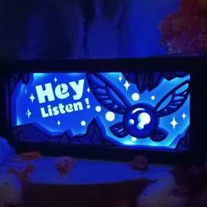 Peut inclure: Un panneau décoratif bleu éclairé avec le texte "Hey Listen!" et un motif de fée stylisé. Le panneau présente un design en couches avec une bordure en forme de grotte et des étoiles, créant un effet lumineux. Le thème général est la fantaisie.