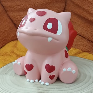 Puede incluir: Una figura de Pokémon rosa y roja con corazones en su cuerpo. La figura está sentada sobre una superficie de madera.
