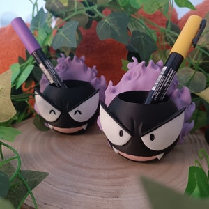 May include: Two black and white cartoon ghost-shaped pencil holders with purple flames. The holders are filled with purple and yellow markers.