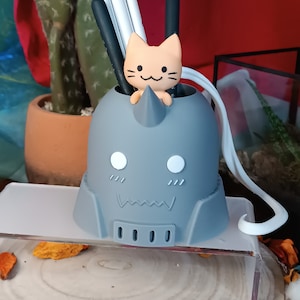 Peut inclure: Organiseur de bureau de style anime gris et blanc en forme de tête de personnage avec une petite figurine de chat orange qui sort du haut.