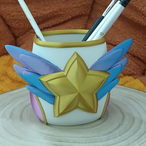 Peut inclure: Un porte-crayons en céramique blanche avec une étoile dorée et des ailes bleues et violettes. Le porte-crayons a une bordure dorée et est posé sur une surface en bois.