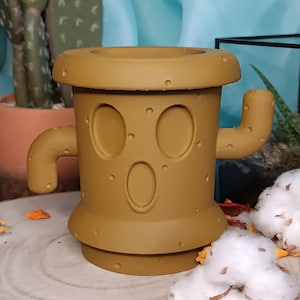 Peut inclure: Un pot de cactus en céramique marron avec un visage et des bras. Le pot est posé sur une surface en bois.