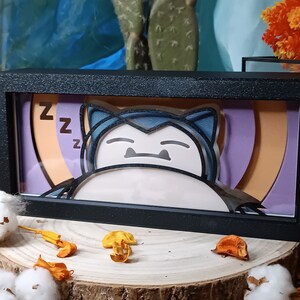 Peut inclure: Une boîte à ombre encadrée de noir avec un Snorlax Pokémon en papier découpé en 3D. L'arrière-plan est un dégradé de couleurs violet, orange et jaune avec les lettres "ZZ" dans le coin supérieur gauche.