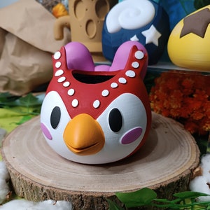 Peut inclure: Un pot de fleurs en forme de hibou rouge et blanc avec un bec et des joues roses. Le pot est décoré de points blancs autour du bord et est posé sur une tranche de bois.