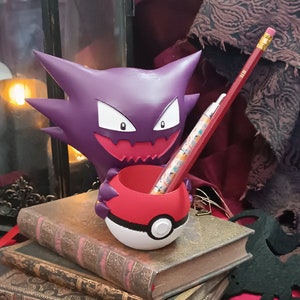 Peut inclure: Une figurine de Gengar de Pokemon, de couleur violette et blanche, tenant un porte-crayons Pokeball rouge et blanc. Le porte-crayons a une bande noire au milieu. Un crayon dépasse du porte-crayons.
