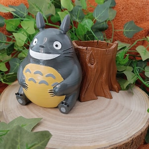 Könnte beinhalten: Eine graue Totoro-Figur mit gelbem Bauch und lächelndem Gesicht sitzt neben einem braunen Baumstumpf-Pflanzgefäß. Die Gegenstände befinden sich auf einer Holzscheibe, umgeben von grünen Blättern und orangefarbenem Stoff.