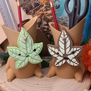 Peut inclure: Deux porte-crayons en céramique brune en forme de personnage de dessin animé avec une feuille verte et brune sur le devant. Le personnage a un visage souriant et une petite tige brune sur le dessus de la tête. Les porte-crayons sont posés sur une surface en bois.